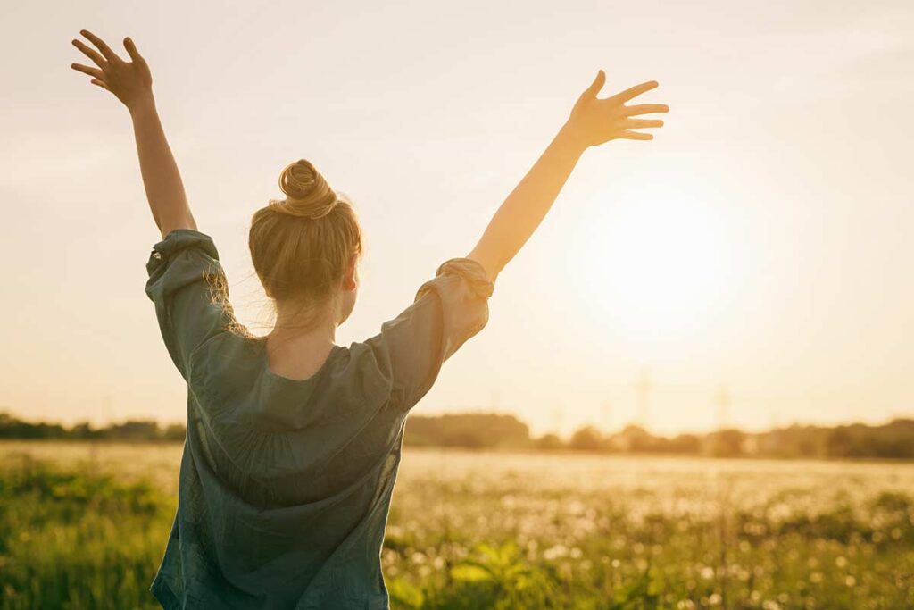 woman facing sunset with two hands up