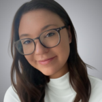 A woman with long brown hair and glasses smiles softly. She is wearing a white turtleneck top and is pictured against a plain, light gray background.