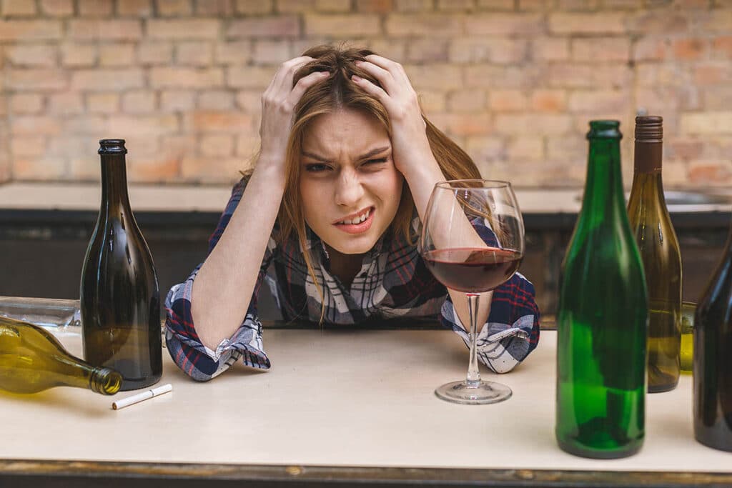 a woman holds her head and wonders am i an alcoholic as she is surrounded by empty wine bottles