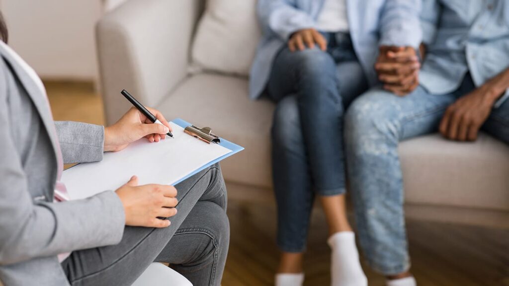 a husband and wife hold hands as they discuss the benefits of marriage counseling with a therapist