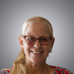 An older woman with long blonde hair, wearing glasses, hoop earrings, and a patterned top, smiles in front of a gray gradient background.
