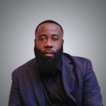A man with a full beard wearing a dark suit and black shirt sits against a plain gray background, looking directly at the camera with a neutral expression.