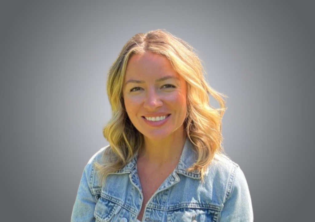 A woman with blonde, wavy hair and a denim jacket smiles at the camera against a plain gray background.