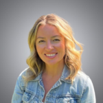 A woman with blonde, wavy hair and a denim jacket smiles at the camera against a plain gray background.