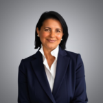 A woman with dark hair, wearing a navy blazer and white blouse, smiles gently at the camera against a plain gray background.